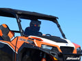A person in a black helmet and goggles drives an orange and white ATV with 1000 and SuperATV branding, equipped with a Polaris General Full Windshield, black roof, and sport seats under a clear blue sky.