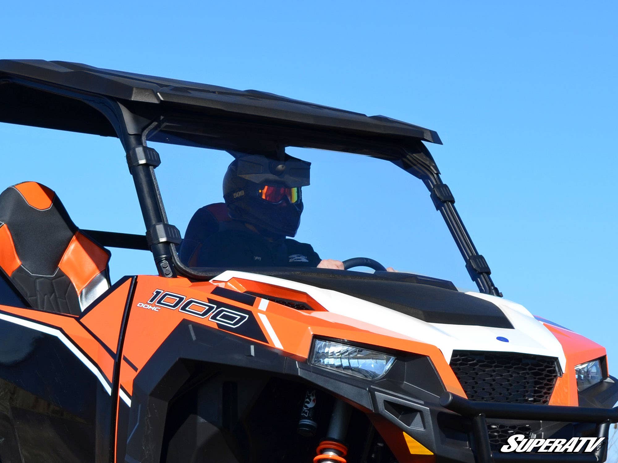 A person in a black helmet and goggles drives an orange and white ATV with 1000 and SuperATV branding, equipped with a Polaris General Full Windshield, black roof, and sport seats under a clear blue sky.