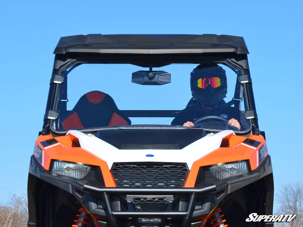 A person wearing a black helmet and goggles drives an orange and white utility vehicle with a protective roof and a Polaris General XP 1000 Full Windshield, viewed from the front against a clear blue sky. SuperATV logo appears at the bottom right.