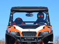 A person wearing a black helmet and goggles drives an orange and white utility vehicle with a protective roof and a Polaris General XP 1000 Full Windshield, viewed from the front against a clear blue sky. SuperATV logo appears at the bottom right.