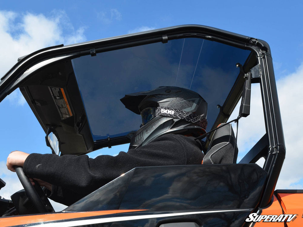 A person in a black helmet and dark clothing sits in the driver’s seat of an off-road vehicle with a clear windshield and a Polaris General Tinted Roof. The blue sky with light clouds reflects, and SuperATV branding appears at the bottom right.