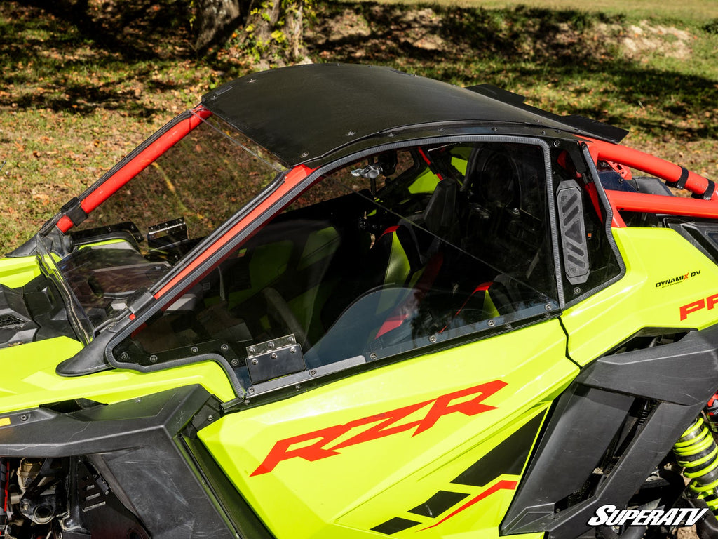 A Polaris RZR Pro R with Hard Cab Enclosure Upper Doors is parked on grass, featuring a bright green and black exterior with red RZR decals, black roof, red roll bars, large windshield, and bucket seats. Sunlit trees appear in the background.