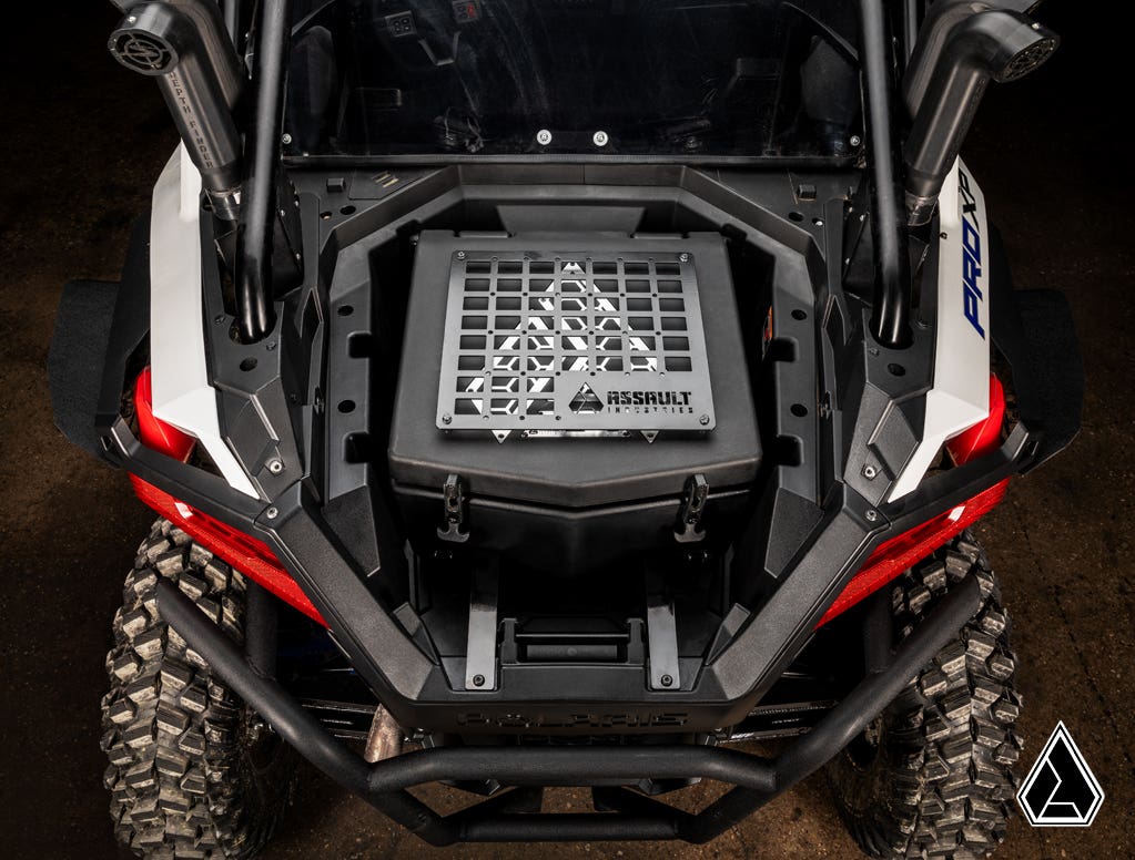 Overhead view of a Polaris RZR PRO XP featuring the Assault Industries Cooler/Cargo Box, mounted in the rear cargo area. The vehicle sports rugged tires, a roll cage, red side panels, and a triangular logo in the bottom right corner.