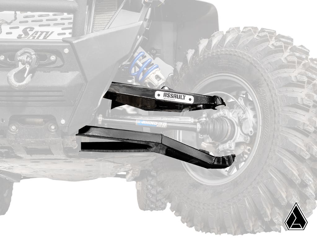 A close-up of a Polaris RZR PRO XP’s suspension featuring Assault Industries High-Clearance 1.5 Forward Offset Boxed A-Arms with a bold “ASSAULT” label, the guard in sharp focus and an angled triangle logo at the bottom right.
