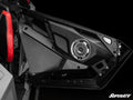 Close-up of Metra 6.5 Speaker Door Pods installed in a Polaris RZR Turbo R. The black door interior features angular, textured panels with a central round speaker. A red and black seat appears on the left, and the SuperATV logo is at the bottom right.