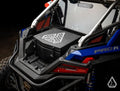 Close-up of the rear cargo area of a blue Polaris RZR Pro R, featuring an Assault Industries Cooler/Cargo Box with a geometric logo secured between metal roll bars. Red taillights, vents, and rugged details are visible against a dark background.