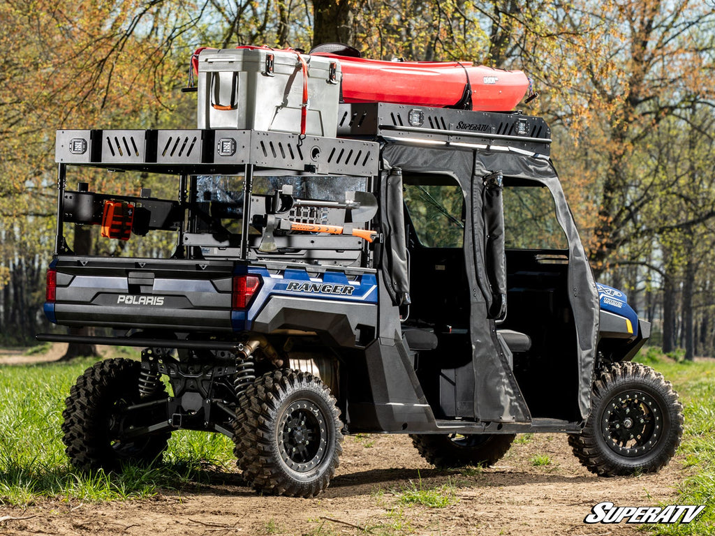 A rugged blue Polaris Ranger outfitted with the XP 570 Outfitter Bed Rack is parked on a dirt path in a grassy, wooded area. It carries a roof-mounted kayak, storage cases, chainsaw, and gear racks for outdoor adventure. The doors are open.