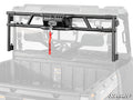 A Polaris Ranger XP 1000 Game Loader Rack—a black metal rack with a winch and red strap—is mounted at the front of a UTV’s cargo bed. The faded vehicle emphasizes the rack, with “SuperATV” branding in the lower right corner.