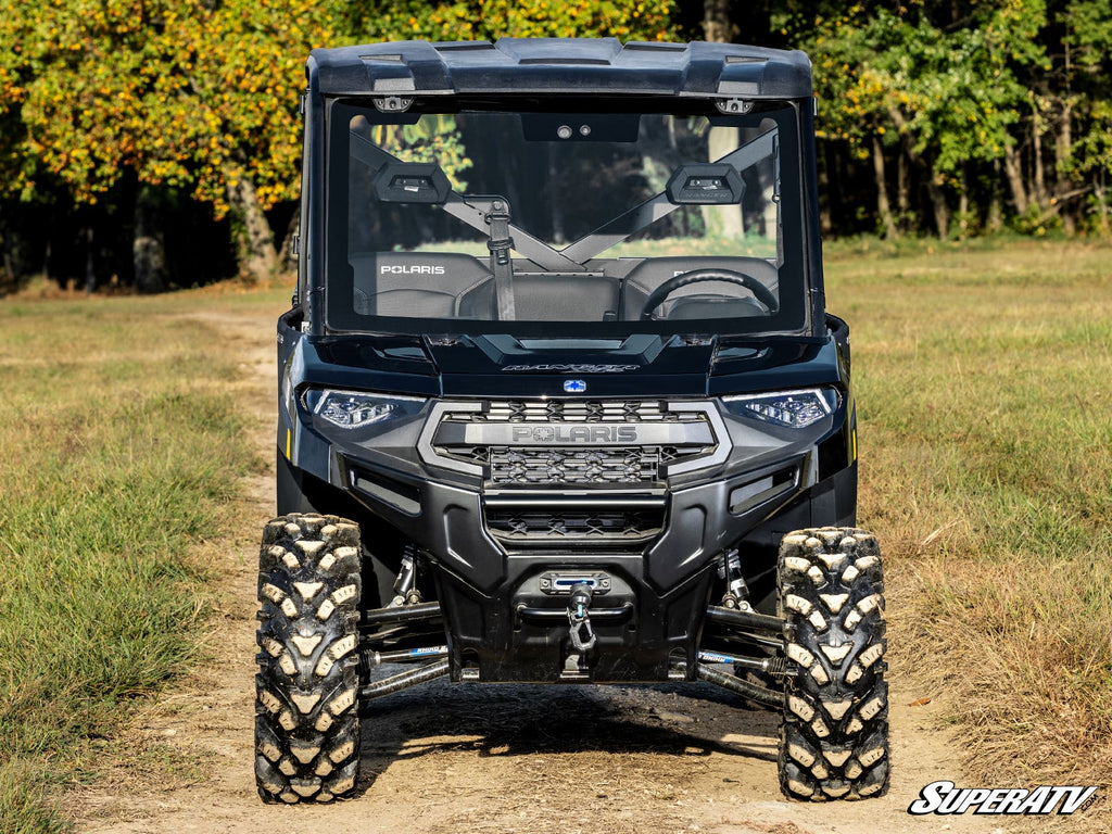 A black Polaris Ranger XP with the Kinetic MaxDrive Power Flip Glass Windshield is parked on a dirt path edged by grass, with trees in the background. The rugged front view features tough tires, a winch, and the Polaris grille logo.