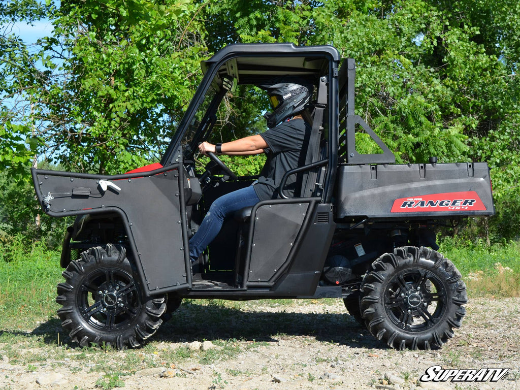A person wearing a helmet sits inside a black Polaris Ranger with Midsize 570 Aluminum Doors and large off-road tires, parked on gravel amid green trees. The driver faces right, and the vehicle’s aluminum door is open.