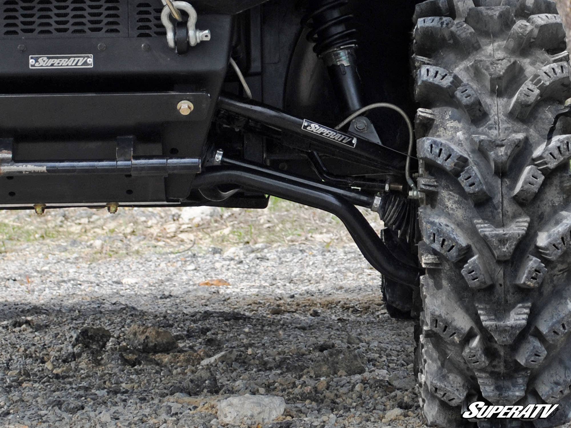 Close-up of a Polaris Ranger 700 with High Clearance 1 Forward Offset A-Arms by SuperATV, showing rugged front suspension and large knobby tire on rocky ground. SuperATV logo is visible in the bottom right corner.