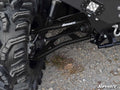 Close-up of a Polaris Ranger XP 900 High Clearance 1.5 Forward Offset A-Arms connecting a large, knobby tire to the chassis on gravel. The SuperATV logo is visible, along with part of the chassis, shock absorber, and an LED light.