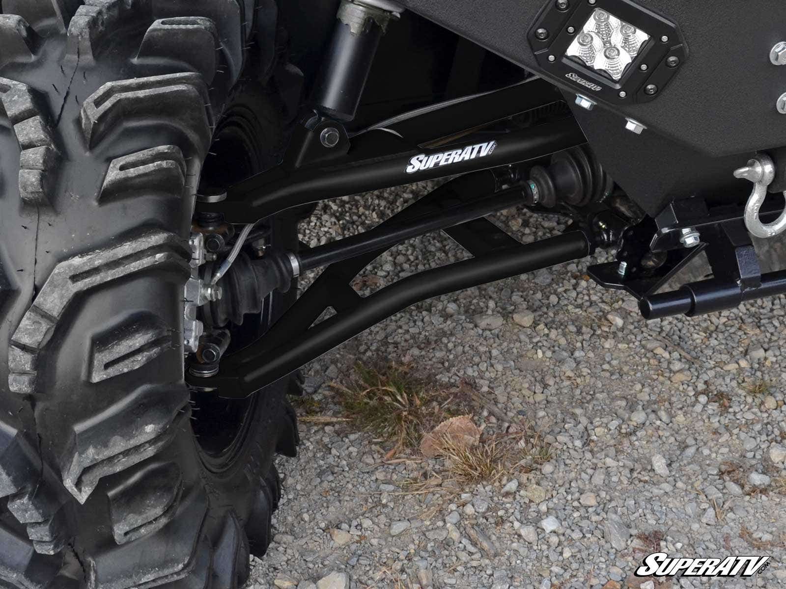 Close-up of a Polaris Ranger XP 900 High Clearance 1.5 Forward Offset A-Arms connecting a large, knobby tire to the chassis on gravel. The SuperATV logo is visible, along with part of the chassis, shock absorber, and an LED light.