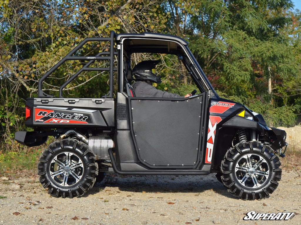 A person in a helmet and dark clothing sits in a black and red Polaris Ranger XP 900 featuring Heavy-Duty Rock Slider Nerf Bars, parked on gravel with green trees behind. The SuperATV logo is visible in the corner.