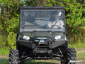 A person wearing a helmet and goggles drives a green off-road utility vehicle with thick tires, a front winch, and a Polaris Ranger 900 Diesel Full Windshield. Green trees and grass surround the vehicle. The SuperATV logo appears in the lower right corner