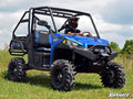 A person wearing a helmet drives a blue Polaris Ranger XP 570 with a 4 Portal Gear Lift over grassy terrain. The vehicle features rugged tires, a roll cage, and black side panels. Trees and a blue sky with clouds are in the background.