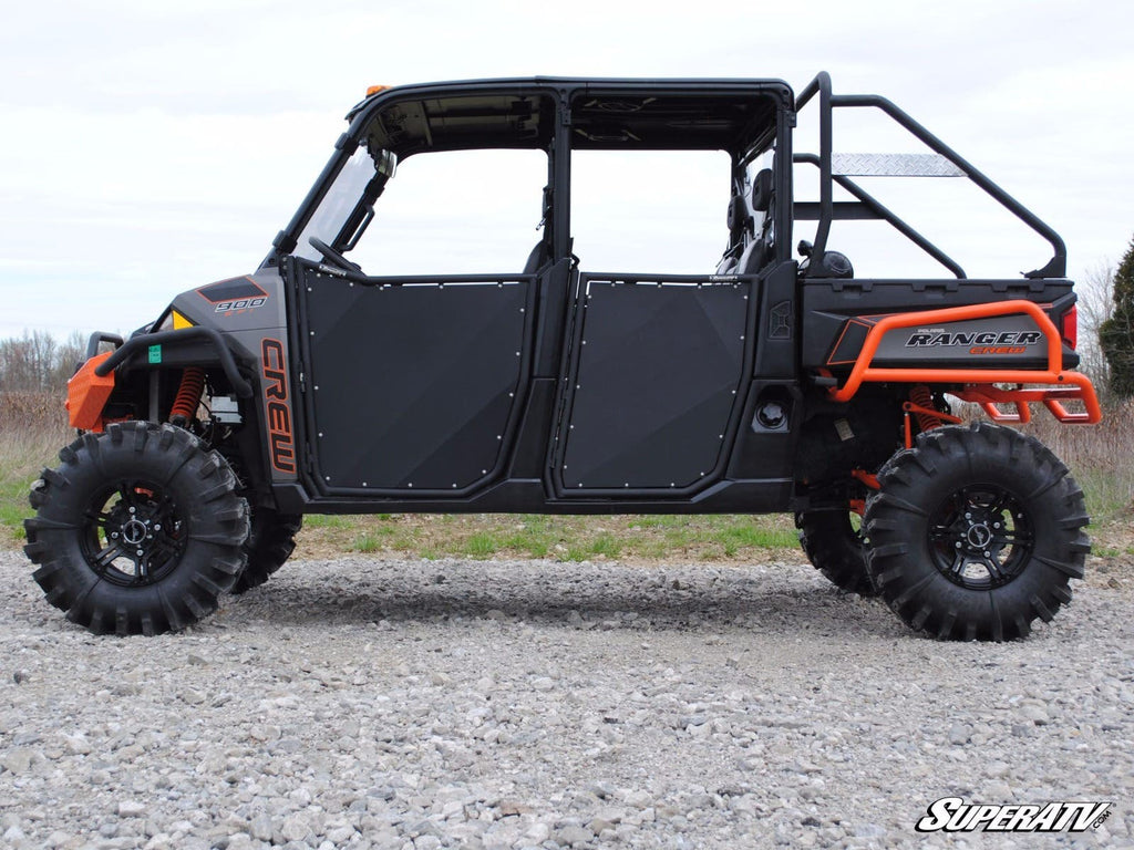 A Polaris Ranger UTV, featuring aluminum lower doors from the Ranger XP 570, has lifted suspension, large off-road tires, a visible roll cage, and seats four. The black and orange vehicle is parked on gravel with trees and sky behind it.