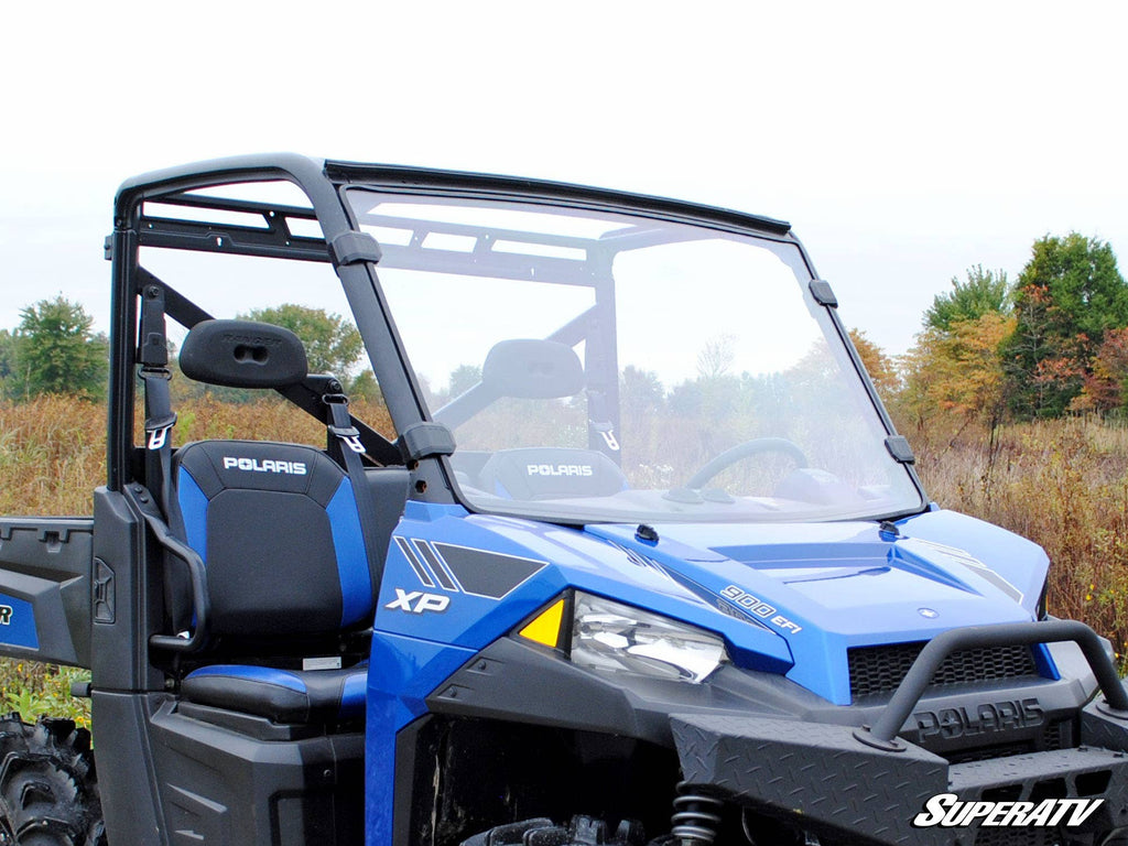 A blue Polaris Ranger XP 570 with a clear full windshield is parked on a grassy field. The vehicle has two black and blue bucket seats, visible SuperATV branding, and trees with green and autumn leaves in the background.
