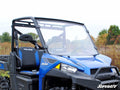A blue Polaris Ranger XP 570 with a clear full windshield is parked on a grassy field. The vehicle has two black and blue bucket seats, visible SuperATV branding, and trees with green and autumn leaves in the background.