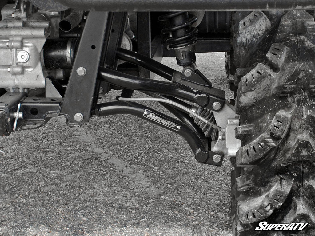 Close-up of Polaris Ranger High Clearance Rear A-Arms’ suspension system with black control arms, visible shock absorber, and rugged tire on gravel. SuperATV branding appears on the A-arm and in the image corner.