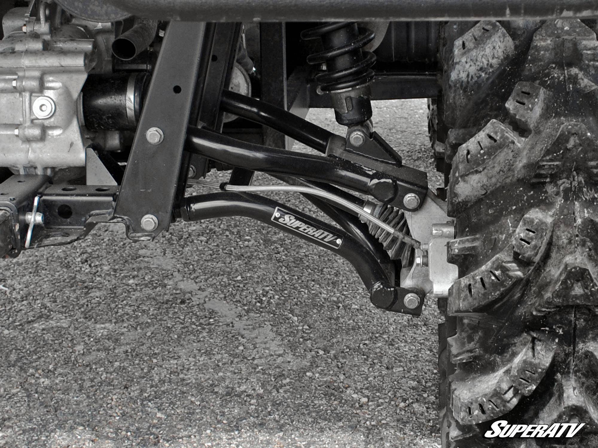 Close-up of Polaris Ranger High Clearance Rear A-Arms’ suspension system with black control arms, visible shock absorber, and rugged tire on gravel. SuperATV branding appears on the A-arm and in the image corner.