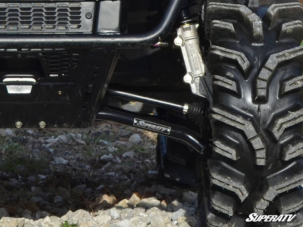 Close-up of a Polaris Ranger Midsize 570 front suspension with High-Clearance 1.5 Forward Offset A-Arms by SuperATV, featuring a large off-road tire, Polaris brake caliper, and the SuperATV logo on rocky terrain.