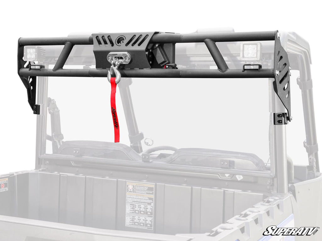 The Polaris Ranger SP 570 Game Loader Rack, a black metal headache rack with built-in winch and red strap, mounts on your UTV bed. It features side brackets, LED lights, the SuperATV logo, and shows a faded UTV cab and bed in the background.