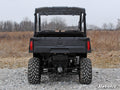 A Polaris Ranger Midsize (2015+) with a rear windshield is parked on rocky ground in an open field with dry grass and leafless trees. The image displays large rugged tires, a flat cargo bed, and the SuperATV logo in the lower right corner.