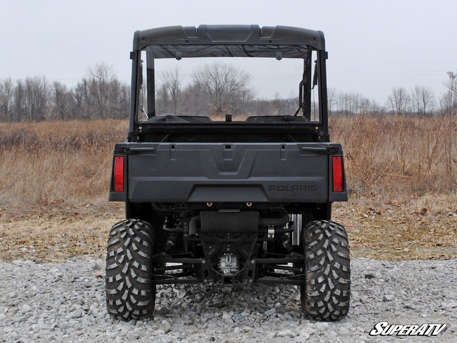 A Polaris Ranger Midsize (2015+) with a rear windshield is parked on rocky ground in an open field with dry grass and leafless trees. The image displays large rugged tires, a flat cargo bed, and the SuperATV logo in the lower right corner.
