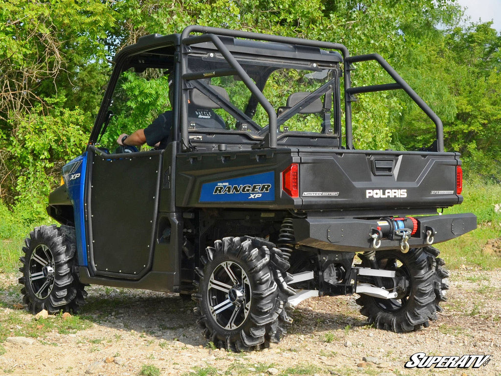 A blue and black Polaris Ranger XP with off-road tires, roll cage, open sides, and cargo bed is parked on a gravel path surrounded by greenery. It features the Polaris Ranger Rear Sport Accessory Bar. The SuperATV logo appears at the bottom right.