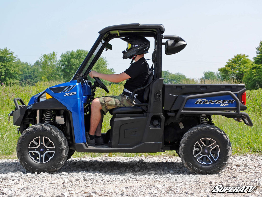 A person in a helmet and black shirt drives a blue Ranger XP on rocky terrain with grass and trees under a clear sky. The Polaris Ranger Wakeboard Speaker Mount is featured, and the SuperATV logo appears in the lower right corner.
