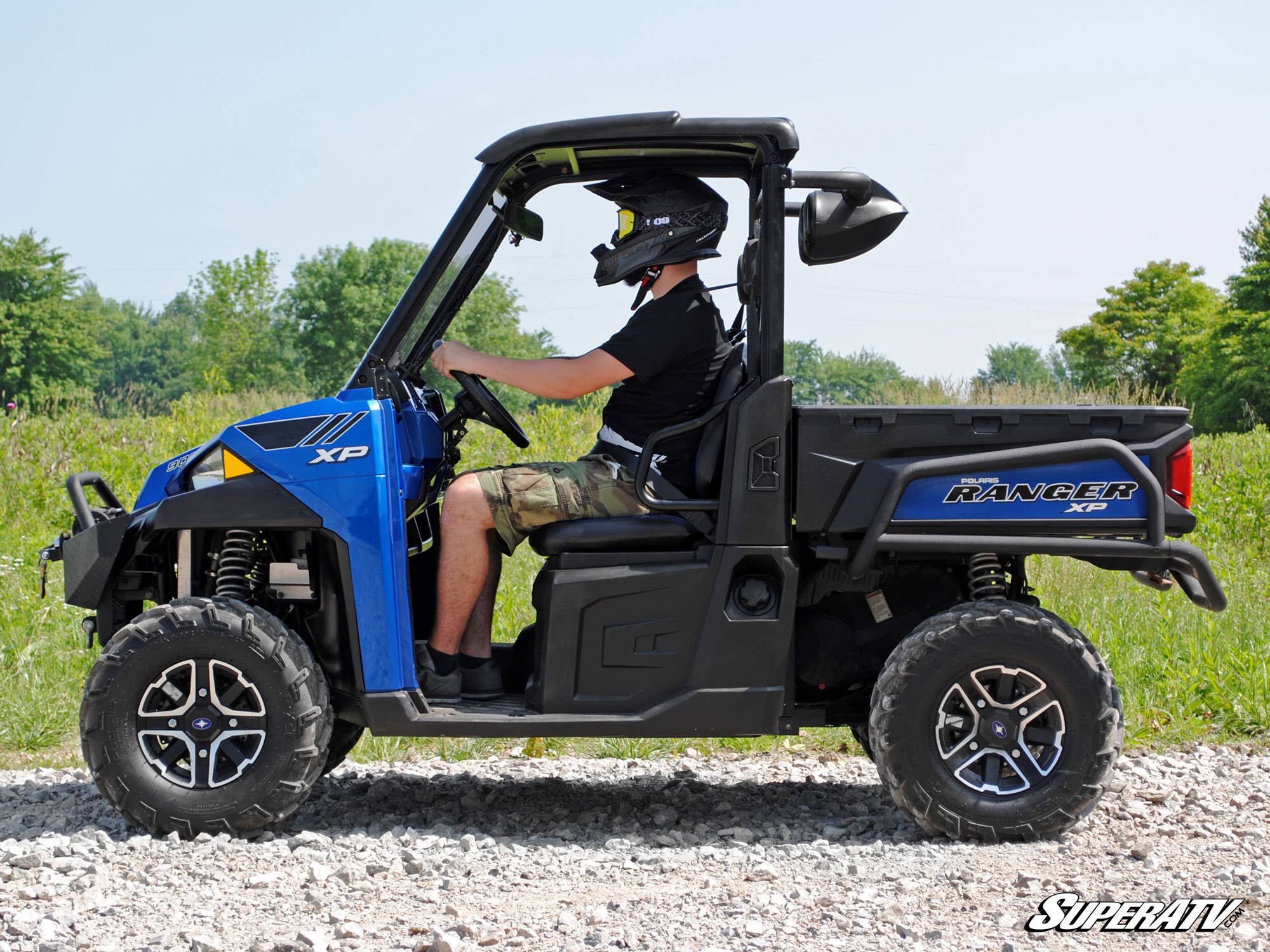 A person in a helmet and black shirt drives a blue Ranger XP on rocky terrain with grass and trees under a clear sky. The Polaris Ranger Wakeboard Speaker Mount is featured, and the SuperATV logo appears in the lower right corner.