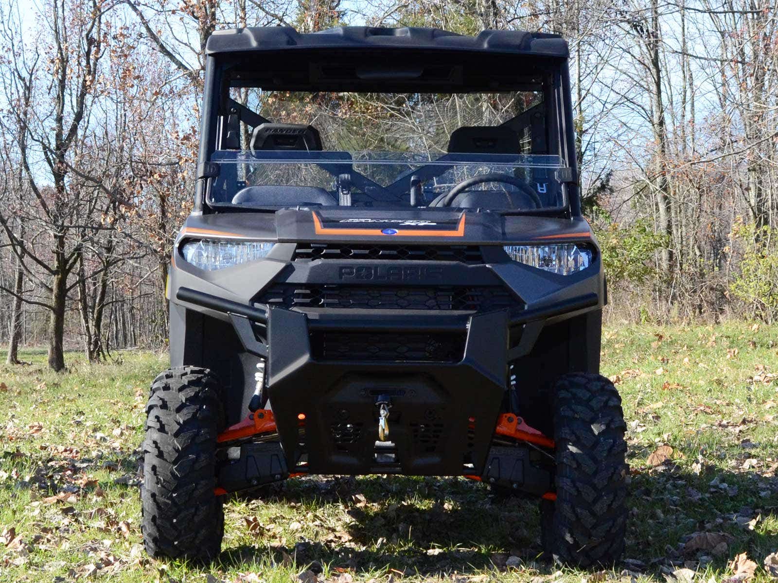 A black utility vehicle with a Polaris Ranger XP 570 Half Windshield and large rugged tires is parked on grass, with trees and dry leaves in the background, hinting at a fall setting.