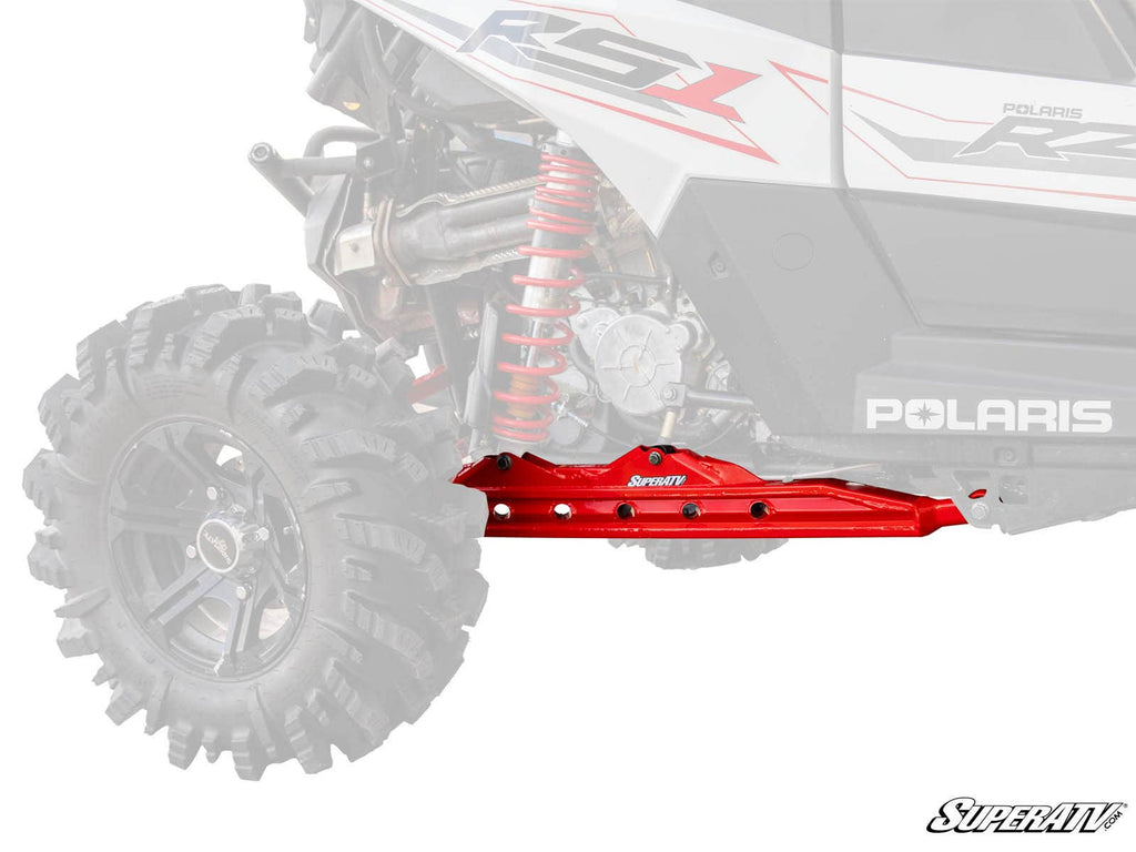 A red Polaris RZR RS1 Rear Trailing Arm is installed on the rear of a Polaris off-road vehicle, standing out against the faded background with its large black tire, visible coil spring, and faint Polaris branding.