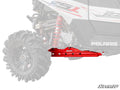 A red Polaris RZR RS1 Rear Trailing Arm is installed on the rear of a Polaris off-road vehicle, standing out against the faded background with its large black tire, visible coil spring, and faint Polaris branding.