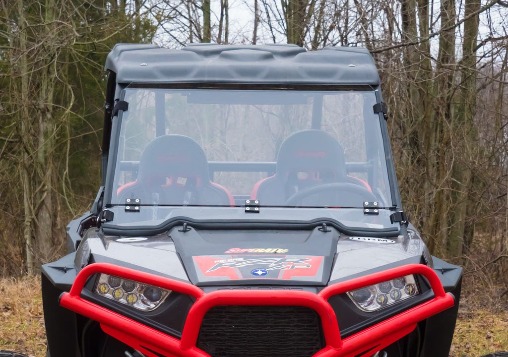 A Polaris RZR S 1000 equipped with a flip down windshield is shown front-facing in black and red, featuring two bucket seats and a detailed dashboard. The background of bare trees and brown grass suggests late autumn or early winter.