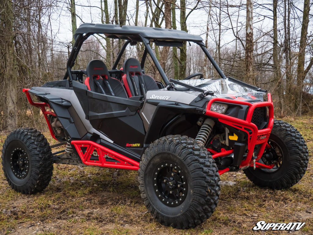 A Polaris RZR XP Turbo with a Flip Down Windshield, featuring black and gray bodywork, red roll bars, large knobby tires, and two bucket seats, sits on muddy ground in a leafless forest. The SuperATV logo is visible in the lower right corner.