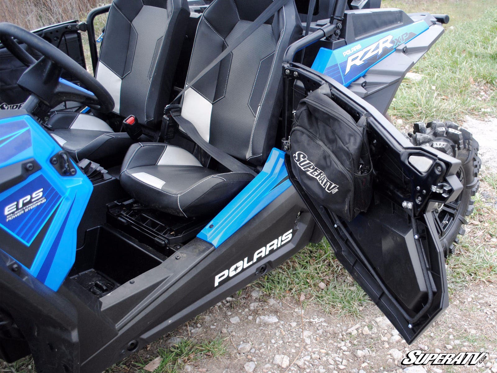 A blue and black Polaris RZR XP 1000 with Full Plastic Doors is parked on rocky ground with grass nearby. The driver-side door is open, showing black seats with geometric patterns and a SuperATV storage bag inside the door.