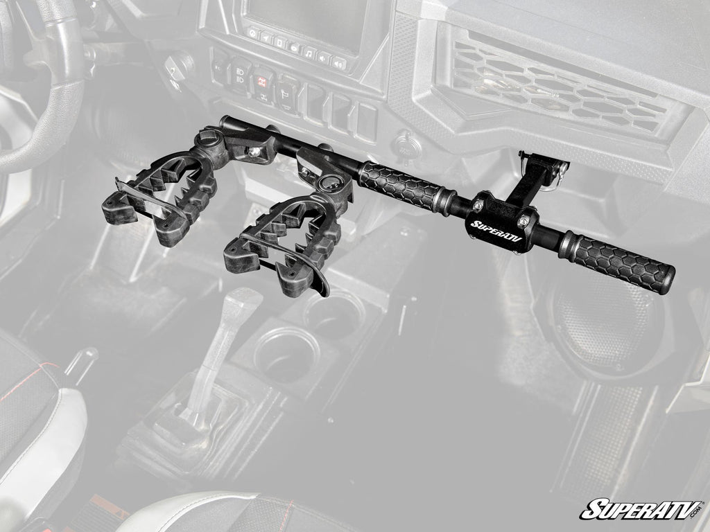 A Polaris RZR Grab Handle Gun Mount is installed between the front seats of a vehicle, holding two guns upright with clamp-style holders and textured grips. The background interior, including seats and gear shift, is partially faded.