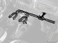 A Polaris RZR Grab Handle Gun Mount is installed between the front seats of a vehicle, holding two guns upright with clamp-style holders and textured grips. The background interior, including seats and gear shift, is partially faded.