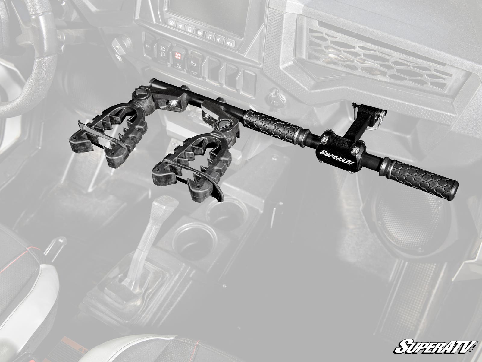 A Polaris RZR Grab Handle Gun Mount is installed between the front seats of a vehicle, holding two guns upright with clamp-style holders and textured grips. The background interior, including seats and gear shift, is partially faded.