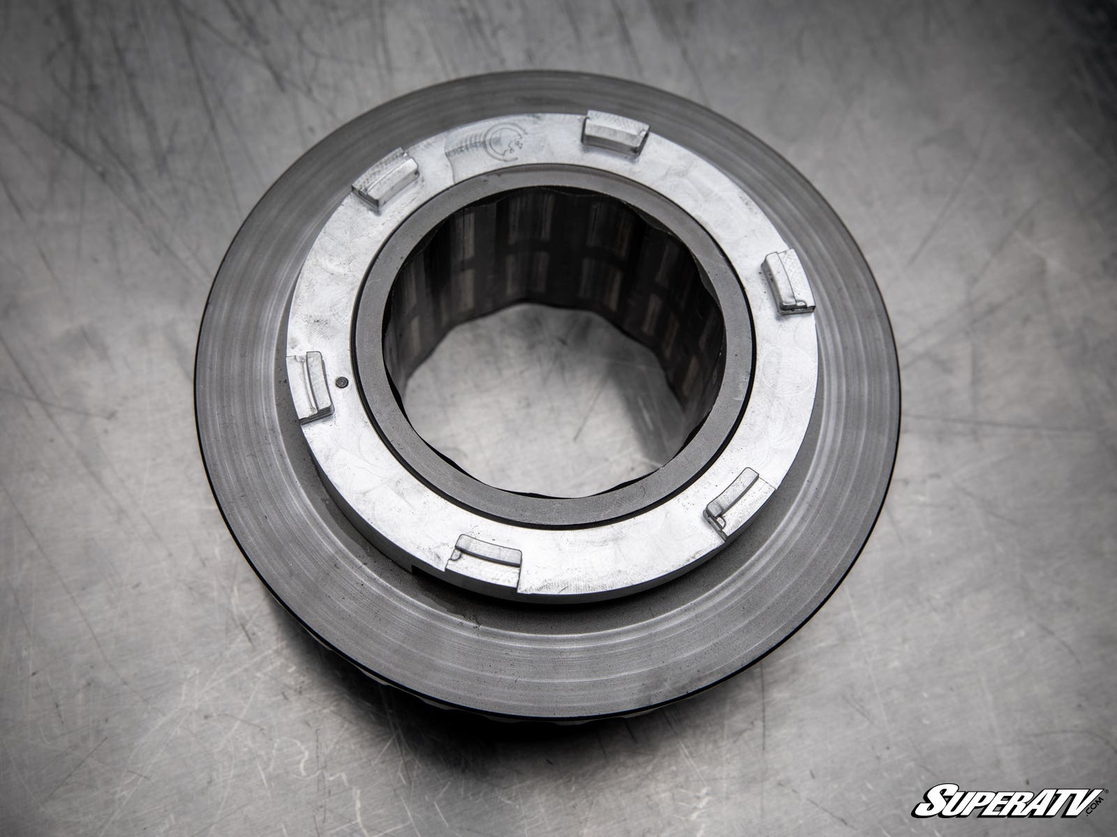 A close-up of a Polaris Ranger Billet Torsion Spring Retainer shows its round shape, grooved edges, and rectangular tabs on a scratched metallic surface. The SuperATV logo is visible in the lower right corner.