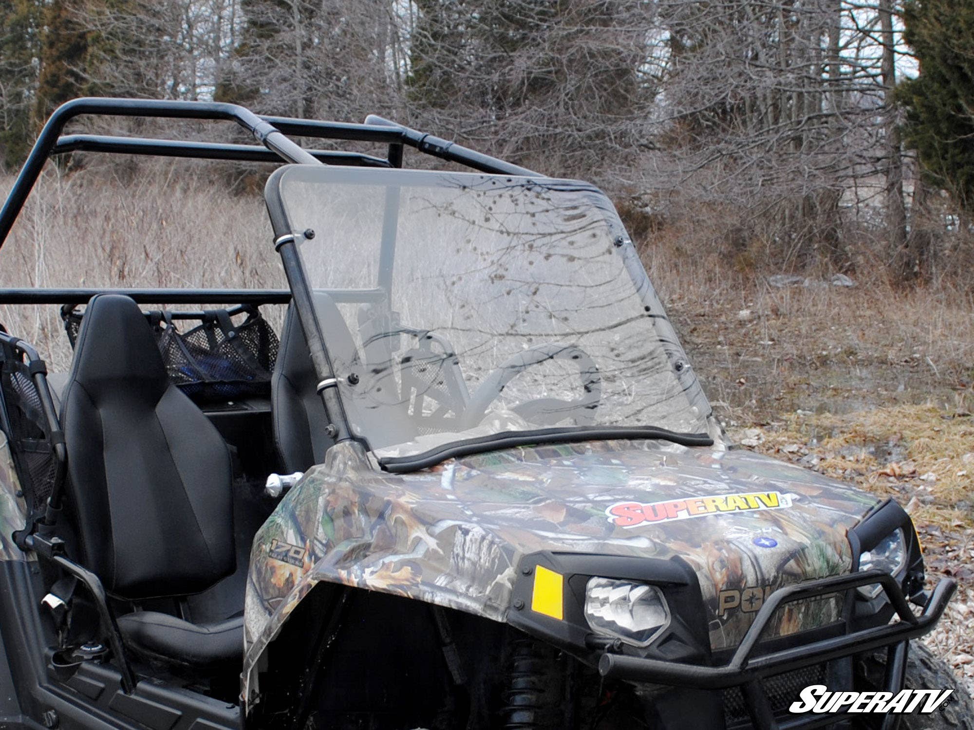 A Polaris RZR 170 with a scratch-resistant full windshield features a camo pattern, two black bucket seats, roll cage, and SuperATV branding. It’s parked outdoors on dry grass near leafless trees.