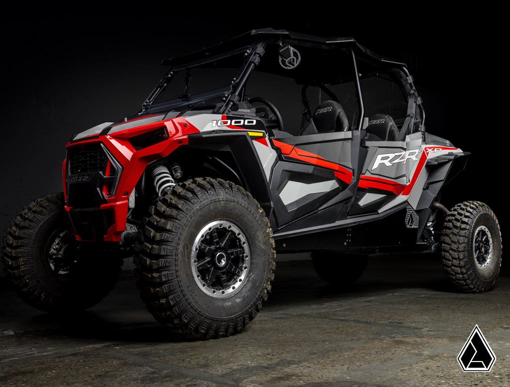 A black and red Polaris RZR XP 4 Turbo with Assault Industries Rock Sliders, bold 1000 and RZR graphics, roll cage, open sides, seating for four, and large knobby tires is shown in a dark studio with a geometric logo at bottom right.