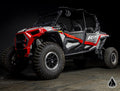 A black and red Polaris RZR XP 4 Turbo with Assault Industries Rock Sliders, bold 1000 and RZR graphics, roll cage, open sides, seating for four, and large knobby tires is shown in a dark studio with a geometric logo at bottom right.