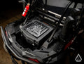 A close-up top view of a Polaris RZR XP Turbo shows the Assault Industries Cooler/Cargo Box with a grid top and geometric logo. Nearby, a bright red fire extinguisher is mounted on the frame, amid protective pipes and bars.