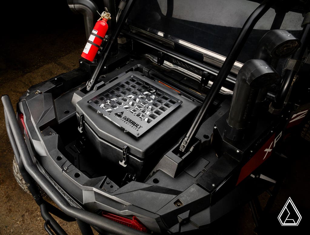 A close-up top view of a Polaris RZR XP Turbo shows the Assault Industries Cooler/Cargo Box with a grid top and geometric logo. Nearby, a bright red fire extinguisher is mounted on the frame, amid protective pipes and bars.