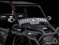 A close-up of a black Assault Industries Polaris RZR XP 1000 Windscreen shows the angular hood, windshield, side mirrors, and RZR-branded seats. The image features a black background and a white geometric logo in the lower right corner.