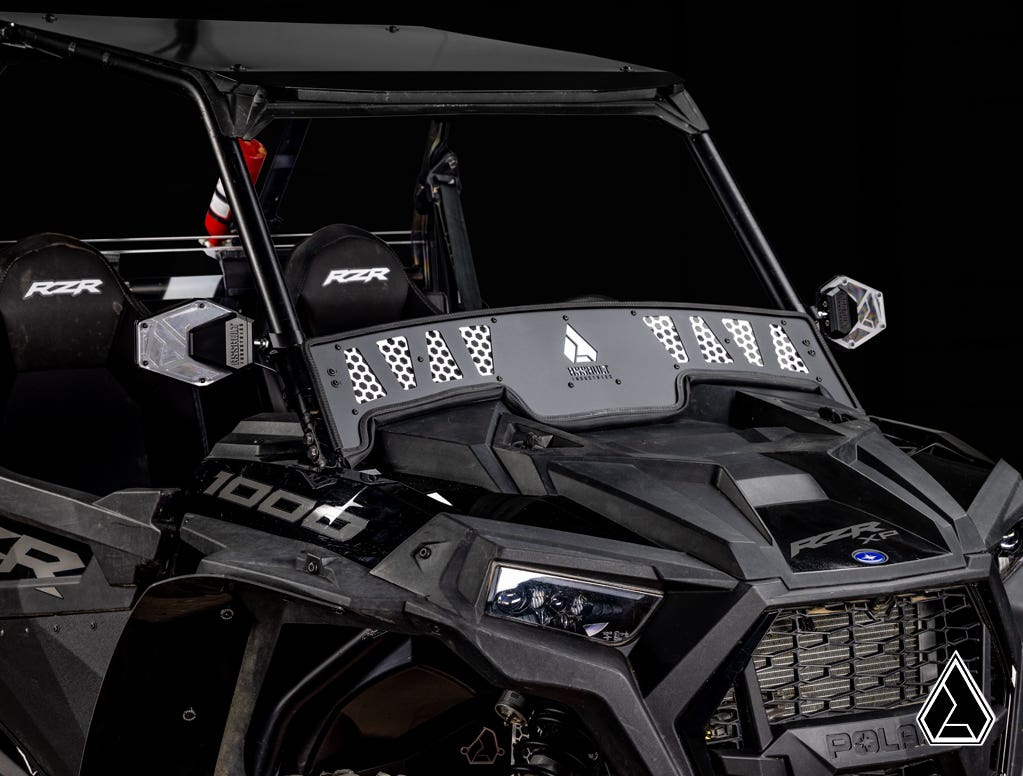 A rugged black Polaris RZR with angular bodywork is shown against a black background, featuring the Assault Industries Polaris RZR XP Turbo Windscreen with a tinted panel, geometric perforations, and visible logo.