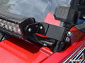 Close-up of a SuperATV Polaris RZR 30 Light Bar Mounting Bracket - Flush to Hood, securing a black LED light bar to a red vehicle. The sturdy mount is attached to the frame, with windshield and dashboard partially visible.